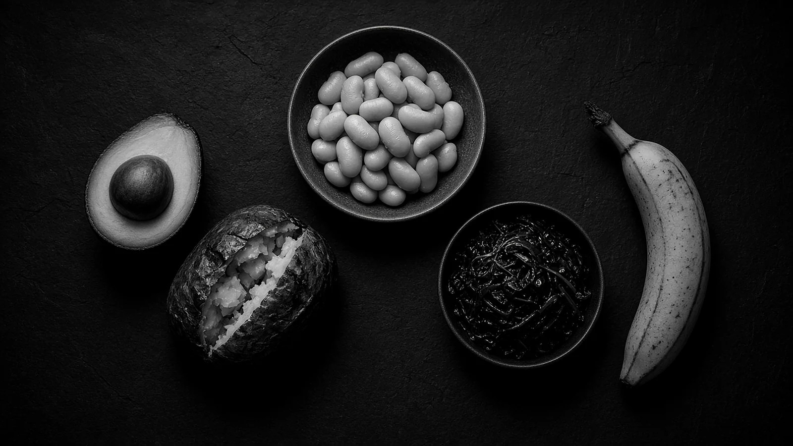 Black-and-white still life of potassium foods: a halved avocado, a baked potato, a bowl of white beans, a bowl of cooked spinach and a banana.